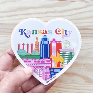 Puede incluir: Una pegatina en forma de coraz&oacute;n con una ilustraci&oacute;n colorida del horizonte de Kansas City. La pegatina incluye el texto "Kansas City" en una fuente arco&iacute;ris. La pegatina est&aacute; sobre una superficie de madera.