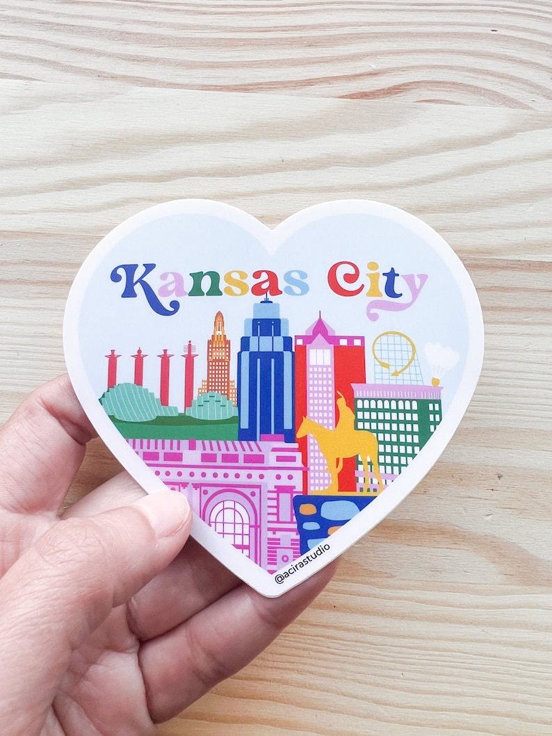 Puede incluir: Una pegatina en forma de coraz&oacute;n con una ilustraci&oacute;n colorida del horizonte de Kansas City. La pegatina incluye el texto "Kansas City" en una fuente arco&iacute;ris. La pegatina est&aacute; sobre una superficie de madera.