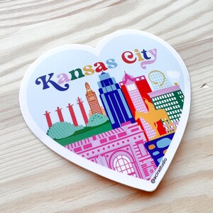 Puede incluir: Una pegatina en forma de coraz&oacute;n con una ilustraci&oacute;n colorida del horizonte de Kansas City. La pegatina presenta edificios, una fuente, un puente y una mula amarilla. El texto "Kansas City" est&aacute; escrito en una fuente arco&iacute;ris en la parte superior de la pegatina. La pegatina tiene un borde blanco y un fondo blanco.