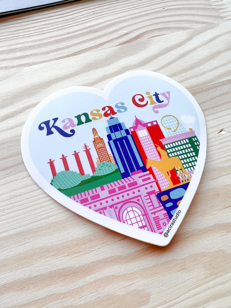 Puede incluir: Una pegatina en forma de coraz&oacute;n con una ilustraci&oacute;n colorida del horizonte de Kansas City. La pegatina presenta edificios, una fuente, un puente y una mula amarilla. El texto "Kansas City" est&aacute; escrito en una fuente arco&iacute;ris en la parte superior de la pegatina. La pegatina tiene un borde blanco y un fondo blanco.