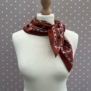 May include: A brown silk scarf with a white and gold paisley pattern. The scarf is tied around a white mannequin's neck.