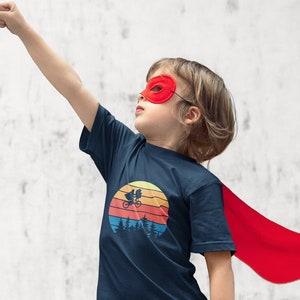 May include: A child wearing a navy blue t-shirt with a retro sunset graphic featuring a bicycle and two figures. The shirt has a red cape and the child is wearing a red mask.