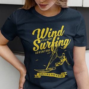 Puede incluir: Una camiseta azul marino con un diseño gráfico amarillo de una persona haciendo windsurf. El gráfico incluye el texto "Wind Surfing" con las palabras "It's all about Adventure" debajo. El gráfico también incluye el texto "Surfers Paradise" en una pancarta.