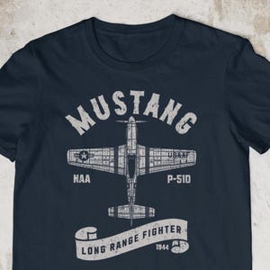 May include: Navy blue t-shirt featuring a white graphic of a P-51D Mustang airplane. The design includes the word "Mustang" above the plane and "Long Range Fighter" below, with "1944" and other text.