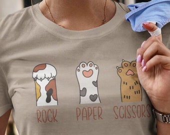 Può includere: Una maglietta marrone chiaro con una grafica di tre zampe di gatto che rappresentano sasso, carta, forbice. Il testo "Sasso Carta Forbice" è stampato sotto le zampe.