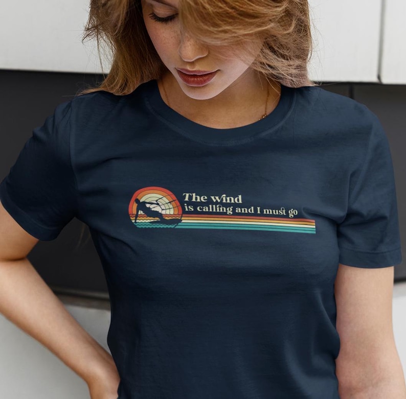 May include: A navy blue t-shirt with a retro-style graphic of a person waterskiing with the text "The wind is calling and I must go".