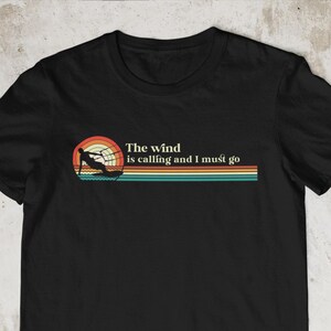 May include: Black t-shirt with a retro-style graphic of a person kitesurfing. The graphic has a sunset color scheme and the text "The wind is calling and I must go".