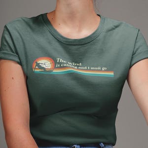 May include: A dark green t-shirt with a retro-style graphic of a person windsurfing. The text "The wind is calling and I must go" is printed on the shirt.