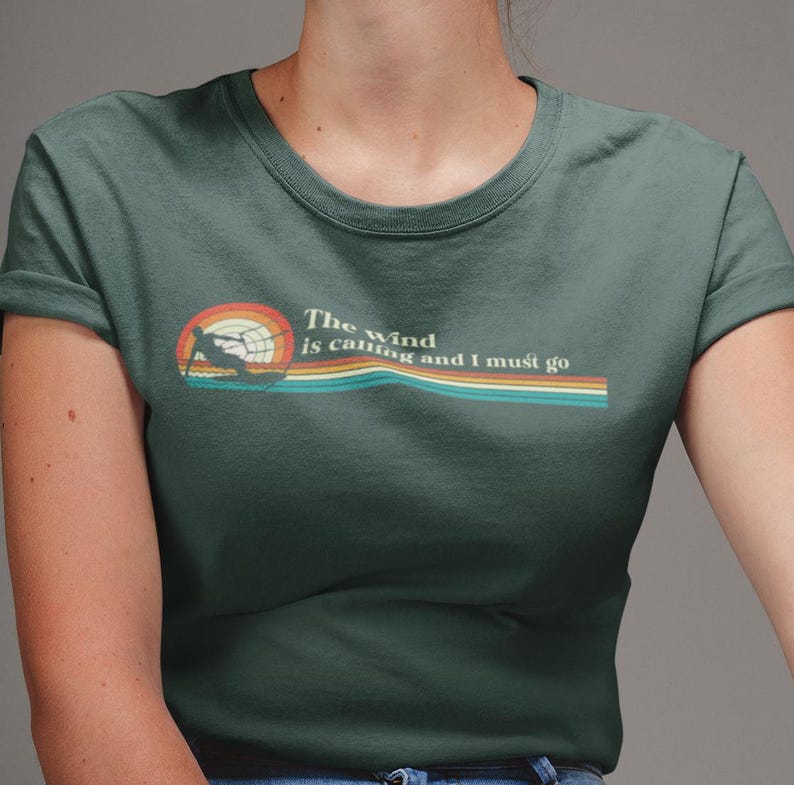 May include: A dark green t-shirt with a retro-style graphic of a person windsurfing. The text "The wind is calling and I must go" is printed on the shirt.