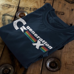 May include: Navy blue t-shirt with the text "Generation X" in a distressed white font and a rainbow stripe design. The shirt is folded on a wooden surface.