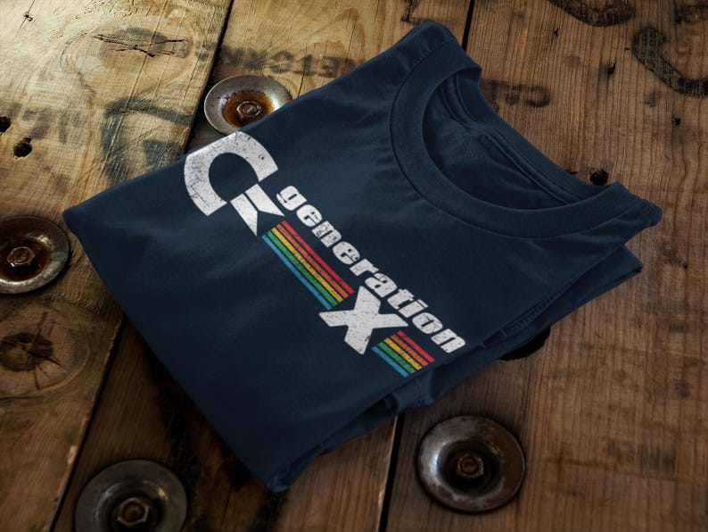 May include: Navy blue t-shirt with the text "Generation X" in a distressed white font and a rainbow stripe design. The shirt is folded on a wooden surface.