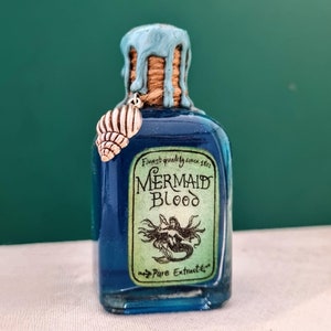 Puede incluir: Una pequeña botella de vidrio azul con un sello de cera azul y blanco y un amuleto de concha de mar plateada. La botella tiene una etiqueta que dice "Finest quality since 1862 Mermaid Blood Pure Extract".