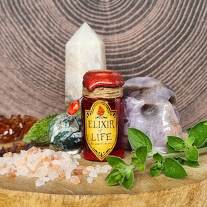 May include: A small red glass bottle with a cork stopper and a yellow label that reads "Elixir of Life" with a red diamond in the corner. The bottle is surrounded by various crystals, a skull, and greenery.
