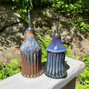 May include: Two ceramic oil bottles with stainless steel spouts. One bottle is brown with blue and bronze accents, the other is blue with teal accents. Both bottles have a ribbed design and are displayed outdoors.