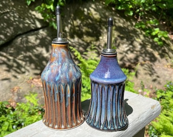 May include: Two ceramic oil bottles with stainless steel spouts. One bottle is brown with blue and bronze accents, the other is blue with teal accents. Both bottles have a ribbed design and are displayed outdoors.
