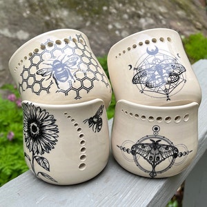 May include: Four ceramic candle holders with black and white designs. The designs feature bees, honeycombs, moths, and geometric patterns. The holders are white with a textured surface.