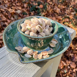 May include: A handmade, teal-colored ceramic serving dish with a small bowl in the center, filled with pistachios. The dish has a leaf-like shape and is placed on a wooden surface, with a blurred background of fall foliage.