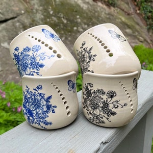 May include: Four ceramic candle holders with a white background and blue floral designs. Each holder has a row of holes around the top edge and a butterfly design.