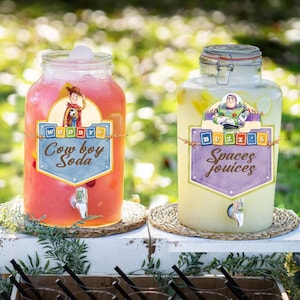May include: Two clear glass beverage dispensers with spigots. The left dispenser contains pink liquid and a label reading Woody's Cowboy Soda. The right dispenser contains yellow liquid and a label reading Buzz's Spaces Jouices.