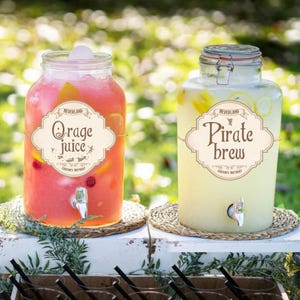 Puede incluir: Dos dispensadores de bebidas de vidrio, uno con "O rage juice" rosa y otro con "Pirate brew" amarillo. Ambos tienen grifos y etiquetas que dicen "Neverland" y "Caron's Birthday". Se encuentran sobre esteras tejidas.