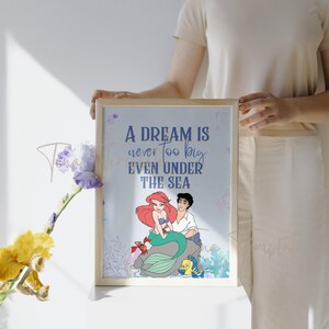 Op de afbeelding: Een blauwe en witte ingelijste print met een citaat dat luidt "Een droom is nooit te groot, zelfs onder de zee". De print toont Ariel en Prins Eric uit de Disney-film De Kleine Zeemeermin.
