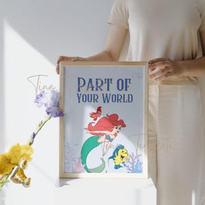 Op de afbeelding: Een ingelijste print met Ariel, de Kleine Zeemeermin, die zwemt met Flounder. De print heeft een blauwe achtergrond met koraal en zeewier. De tekst "Part of Your World" is gedrukt in blauwe letters.