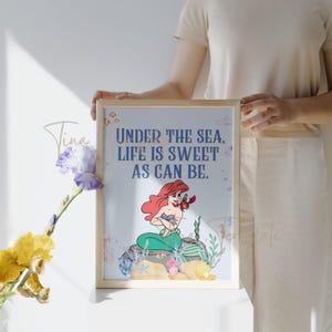 Op de afbeelding: Een lichtblauwe printbare kunstprint met een citaat uit de Disney-film De Kleine Zeemeermin. Het citaat luidt "Onder de zee is het leven zo zoet als het kan zijn." De print toont een cartoonbeeld van Ariel, de zeemeermin, met haar rode haar en haar groene staart. Ze zit op een rots met Sebastian de krab naast haar.