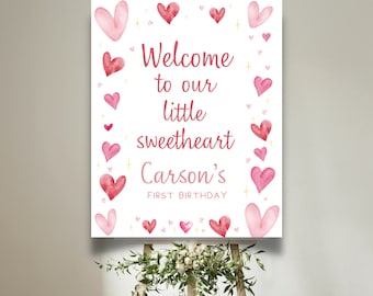 May include: White welcome sign with pink and red watercolor hearts and gold glitter. The sign reads "Welcome to our little sweetheart Carson's First Birthday".
