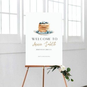May include: A white sign on a wooden easel with the text "WELCOME TO Annie Smith BIRTHDAY BRUNCH". The sign features a watercolour illustration of pancakes with blueberries on a plate. Decorative floral arrangement at the base.