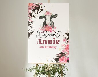 Peut inclure: Une pancarte de bienvenue pour le 4ème anniversaire d'Annie avec une vache noire et blanche portant une couronne de fleurs roses. La pancarte a un fond blanc avec des fleurs roses et des éclaboussures de peinture noire.