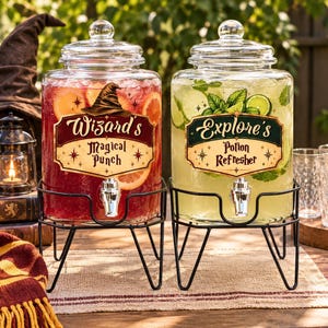 May include: Two clear glass beverage dispensers with silver spigots on black metal stands. The left dispenser contains red liquid with orange slices and a label reading Wizard's Magical Punch. The right contains green liquid with mint and a label reading Explore's Potion Refresher.