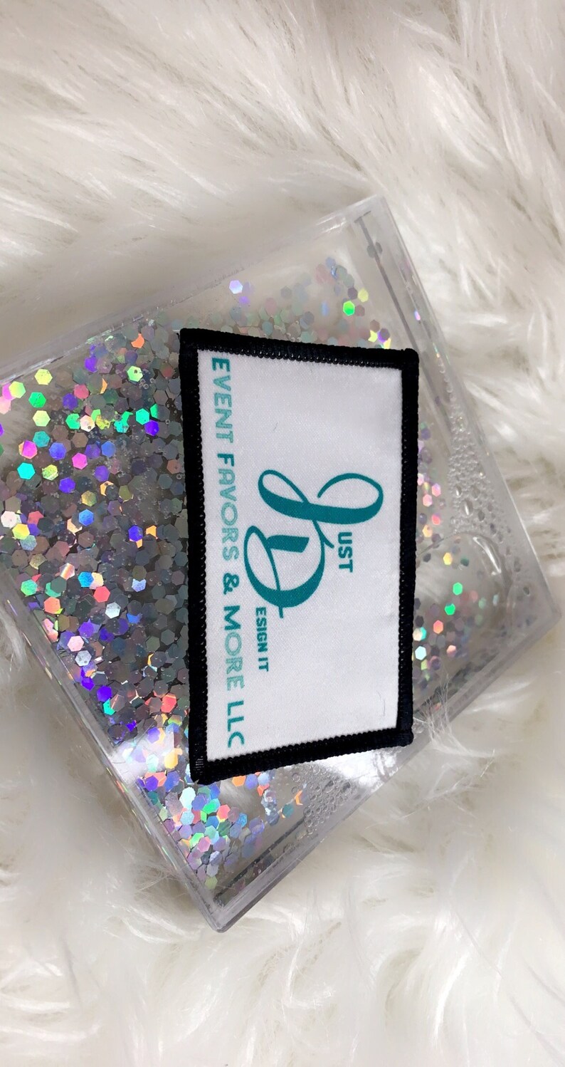 May include: A white square patch with black trim and a teal cursive "D" with the text "Just Design It" and "Event Favors & More LLC" printed on it. The patch is on top of a clear container filled with iridescent glitter.
