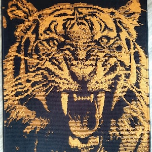 Op de afbeelding: Een close-up van een tijgergezicht, weergegeven in oranje en zwarte gepixelde borduurwerk. De bek van de tijger is open, met scherpe tanden zichtbaar, en zijn ogen zijn intens.