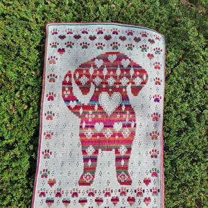 Op de afbeelding: Een rechthoekige, handgehaakte deken met een kleurrijk hondenontwerp. De hond is voornamelijk rood, roze en oranje, met een hart in het midden. De deken heeft een witte achtergrond met pootafdrukken en decoratieve patronen.