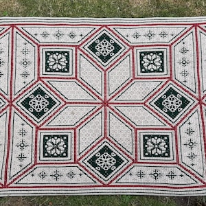 Op de afbeelding: Een handgemaakte gehaakte deken met een geometrisch ontwerp. De deken heeft een crèmekleurige achtergrond met een herhalend patroon van rode en donkergroene geometrische vormen. De deken is rechthoekig en lijkt van zacht garen te zijn gemaakt.