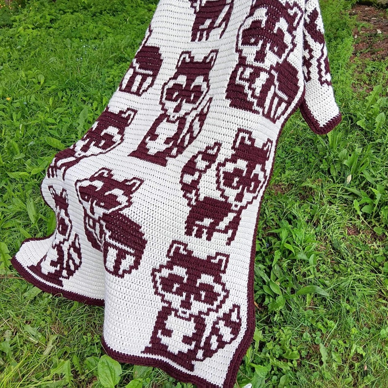 May include: A white crocheted blanket with a brown pixelated raccoon design. The blanket is made with a soft, textured yarn and features a repeating pattern of raccoons.
