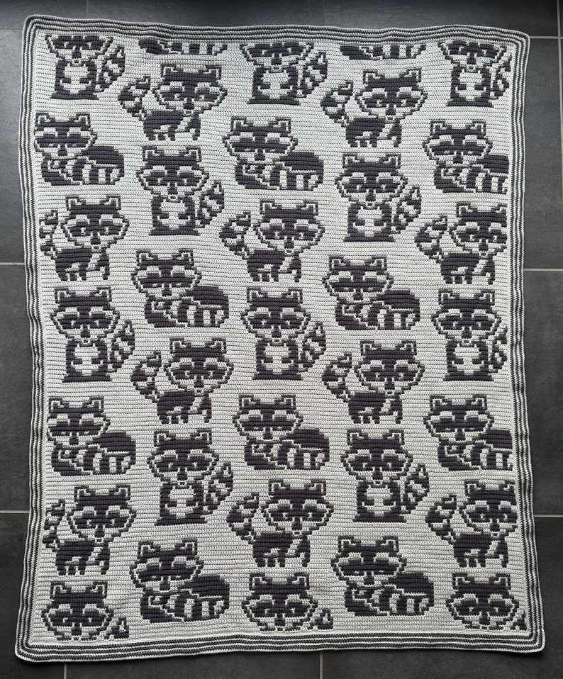 May include: A crocheted blanket featuring a repeating pattern of black and white raccoons. The blanket is made with a soft, textured yarn and has a simple, modern design.