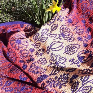 May include: A crocheted throw blanket with a colorful floral pattern in shades of red, purple, and cream. The blanket is laid out on a bed of green grass with yellow daffodils.