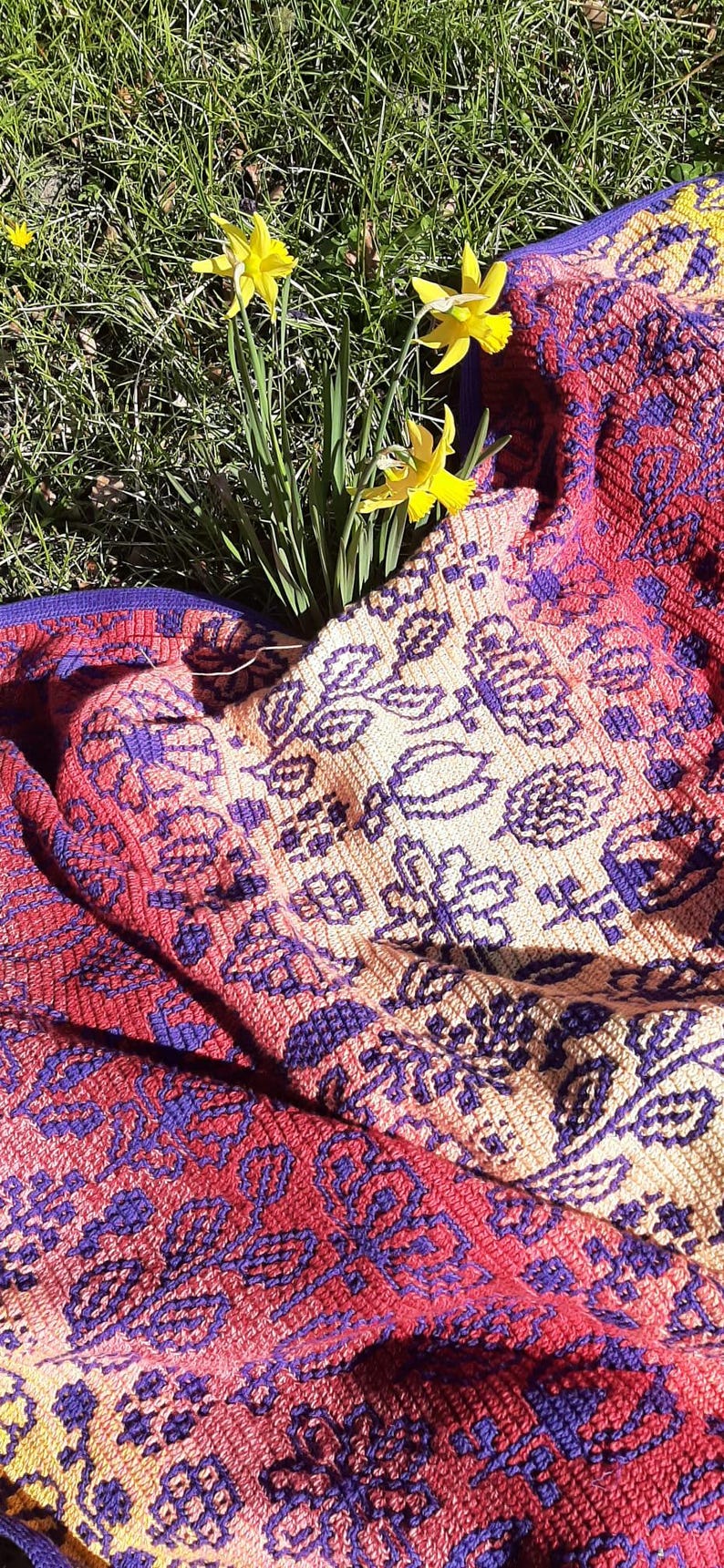 May include: A crocheted throw blanket with a colorful floral pattern in shades of red, purple, and cream. The blanket is laid out on a bed of green grass with yellow daffodils.