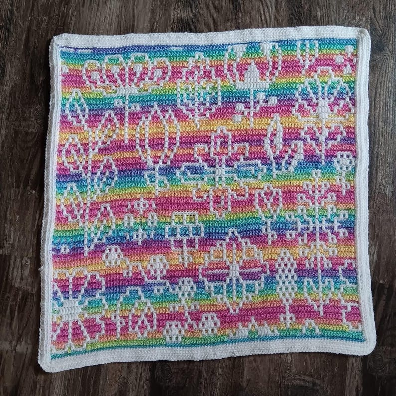 May include: A rainbow crochet blanket with a white border and a floral pattern. The blanket is made with a variety of colors, including pink, blue, green, and yellow.