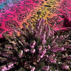 May include: A close-up of a pink and yellow patterned crochet rug with a bush of pink heather flowers in the foreground.