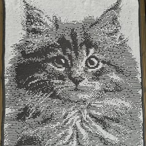 Op de afbeelding: Een close-up van een gehaakt kattenkleed. Het kleed is gemaakt van wit en grijs garen en heeft een gedetailleerd beeld van een kattenkop.