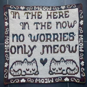 Op de afbeelding: Een crèmekleurige gehaakte deken met de woorden "IN THE HERE IN THE NOW NO WORRIES ONLY MEOW" in donkergroen, blauw en bruin. Twee katten silhouetten en een hart staan onderaan. De rand luidt "MEOW".