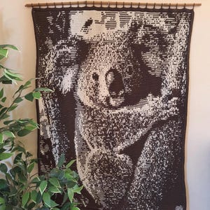 May include: A brown and white crocheted wall hanging featuring a koala bear. The koala is depicted in a pixelated style, with brown and white yarn used to create the image.