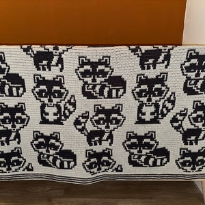 May include: A crocheted blanket featuring a repeating pattern of black and white raccoons on a light gray background.