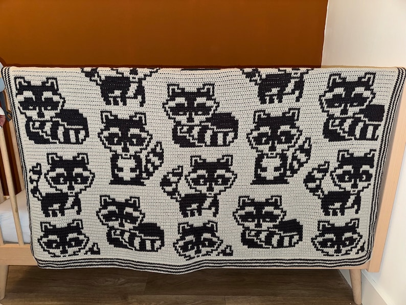 May include: A crocheted blanket featuring a repeating pattern of black and white raccoons on a light gray background.