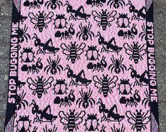 May include: A pink and black crochet blanket with the text "STOP BUGGING ME" repeated around the border. The blanket features a repeating pattern of bees, ladybugs, ants, and grasshoppers.