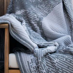 Puede incluir: Una manta de ganchillo gris y blanca sobre una silla de madera. La manta presenta un patr&oacute;n geom&eacute;trico con diferentes texturas y tonos de gris. La manta est&aacute; doblada, mostrando el intrincado trabajo de puntadas y el dise&ntilde;o.