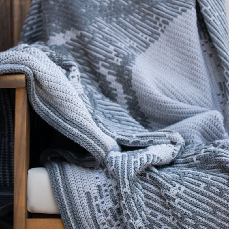 Puede incluir: Una manta de ganchillo gris y blanca sobre una silla de madera. La manta presenta un patr&oacute;n geom&eacute;trico con diferentes texturas y tonos de gris. La manta est&aacute; doblada, mostrando el intrincado trabajo de puntadas y el dise&ntilde;o.