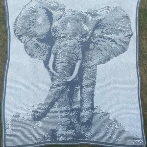 Puede incluir: Una manta de ganchillo con la imagen de un elefante en tonos grises y blancos. El dise&ntilde;o del elefante es detallado, con la trompa y los colmillos bien visibles. La manta tiene un borde gris y est&aacute; colocada sobre hierba. Dimensiones: aprox. 150 cm x 150 cm.